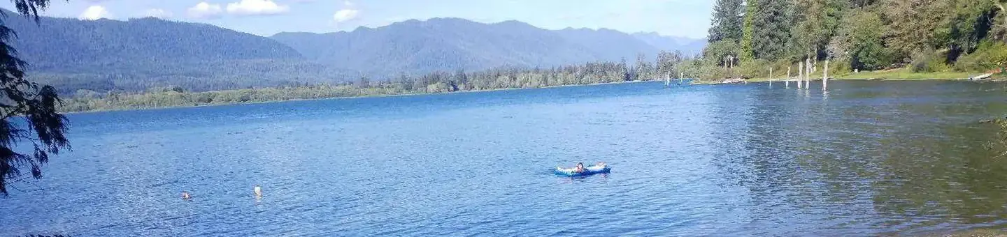 A person on an inflatable raft in a sunlit lake and mountains under a few white clouds floating in blue sky.