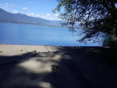 Shaded boat ramp leading to blue, sunlit lake with mountains and clouds floating in blue sky as backdrop.