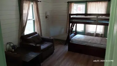 Bunk beds and chairs in the main living room seen from the kitchen. The scene is dimly lit as sunlight enters the room through 3 large windows.
