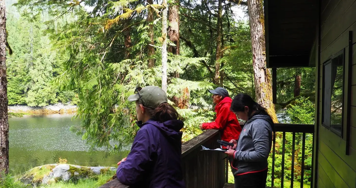 Three people on the patio looking ay the view from Frosty Bay Cabin with trees and water