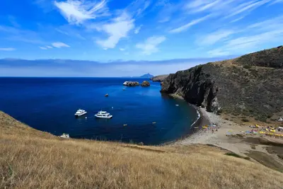 Harbor with boats, kayaks, and visitors on the beach. Steep island cliffs and other islands in the distance.