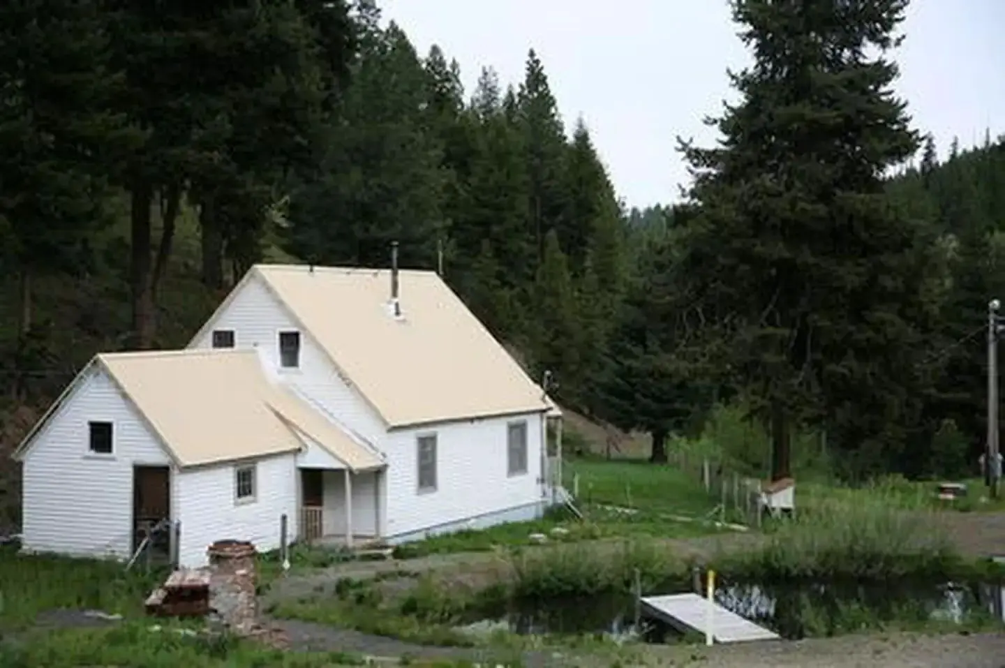 White house on flat green grass between a conifer covered hill and a small pond with a dock.