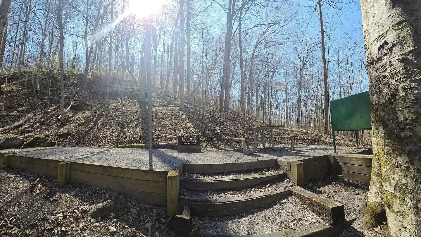 The sun shines down onto a gravel tent pad that has a picnic table and grill.