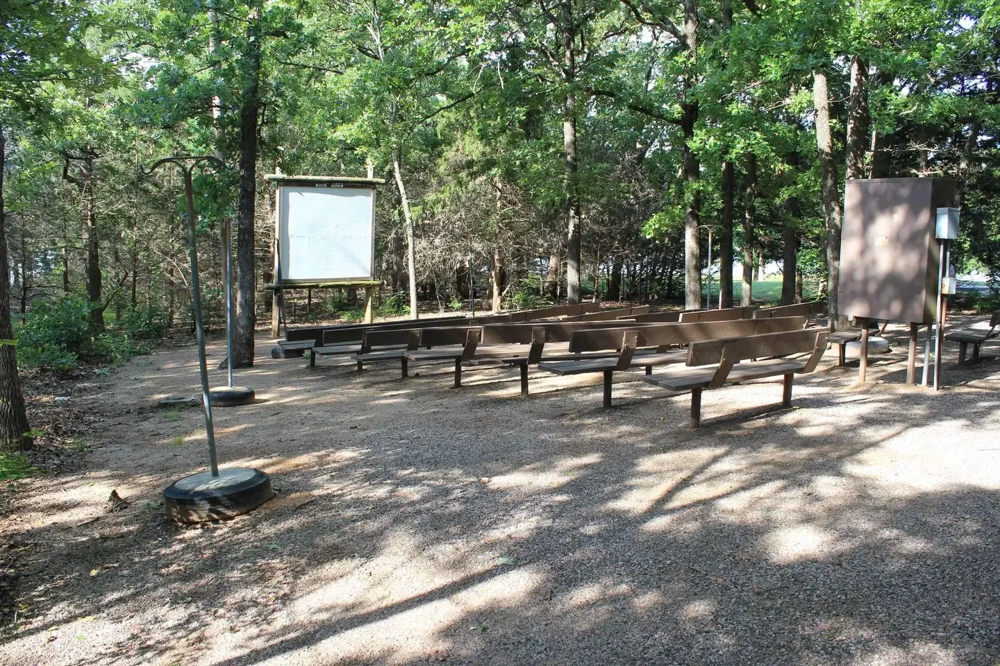 surrounded by trees, an ampitheater with wooden benches faces away from the viewer. A white projector board is in front of the benches.