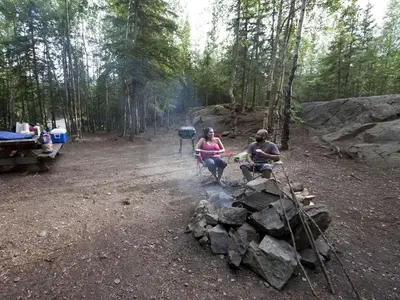 Campers relax in campground next to smoking campfire, background of forest and a picnic table holding supplies.