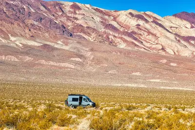 Gray campervan on roadside campsite with green brush vegetation in the foreground and dramatic reddish and white stratified mountains in the far background.