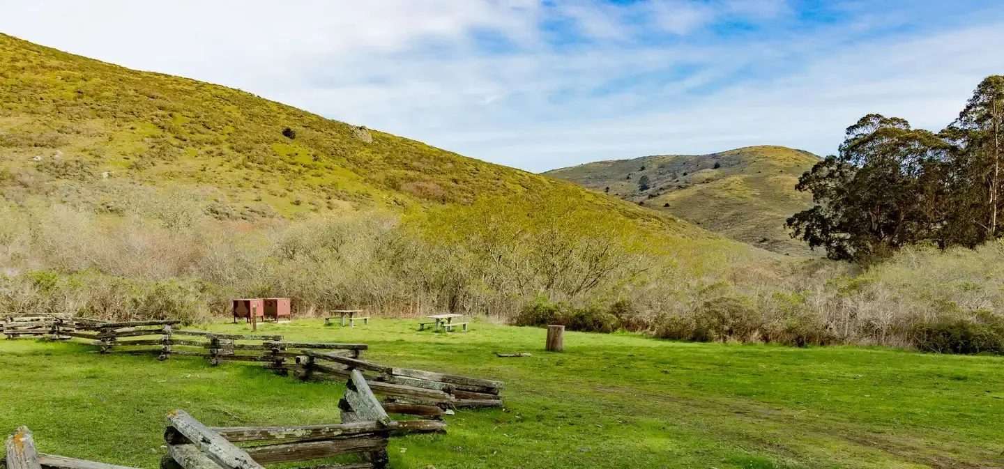 An image of a grassy meadow 2 picnic tables and bear lockers, and green hills in the distance.