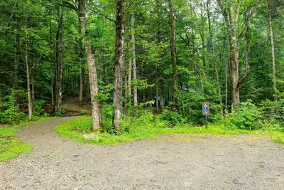 A gravel parking lot in front of a green and shaded campground. An ADA parking space is in front of the campground.
