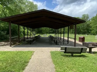 A view of an open sided pavilion with picnic tables, pedestal BBQ grill, and benches with a background of green vegetation.