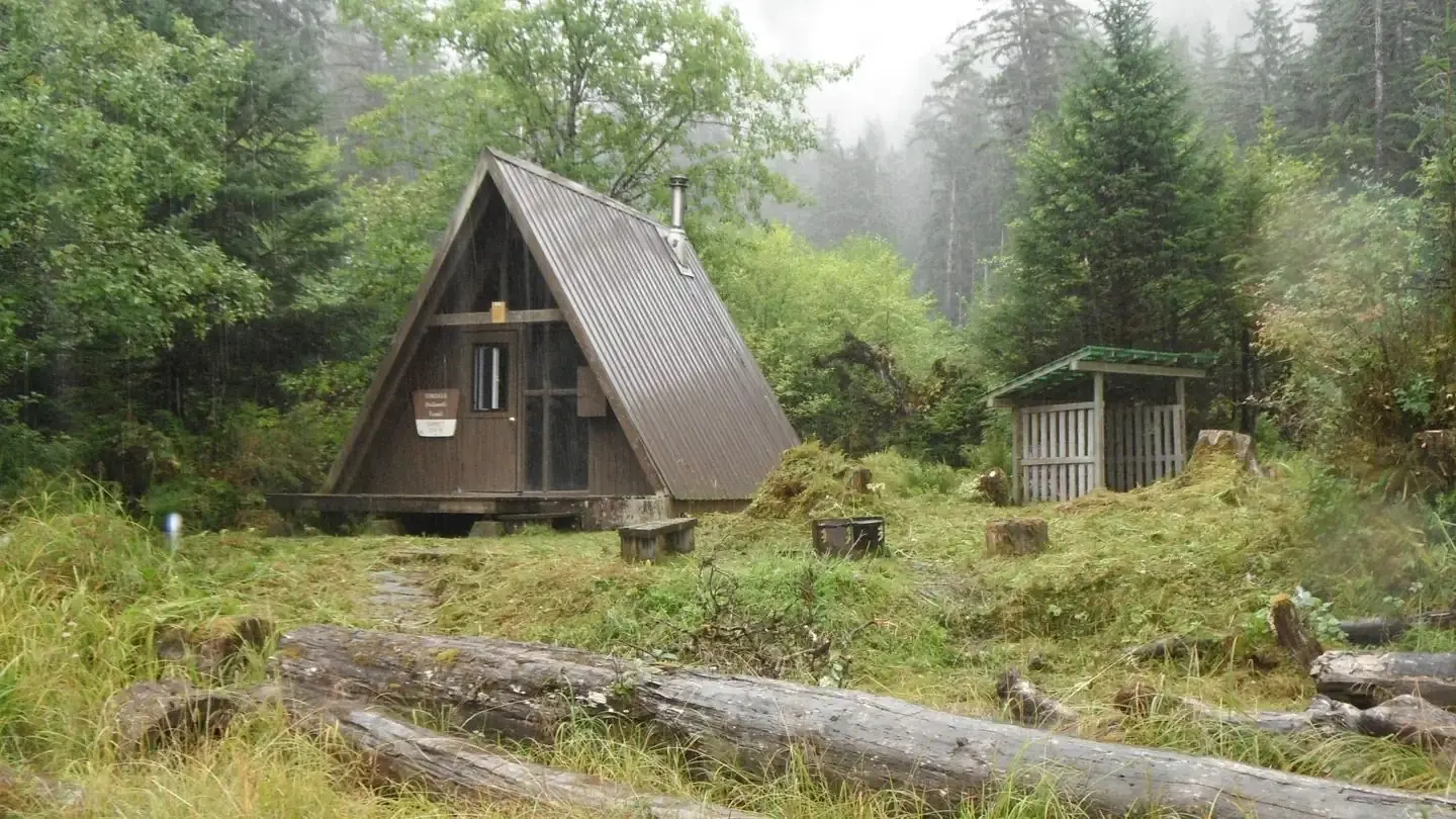 Brown A Frame Cabin, Garnet Ledge, with structure for stacked wood with trees around and two downed logs in front of cabin