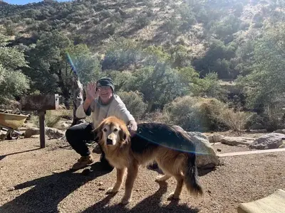 A man squatting down next to his dog, the man is smiling and waving at the camera.