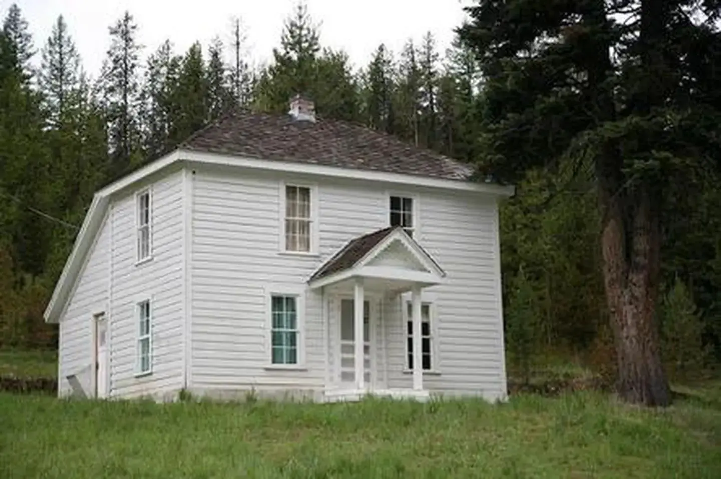 White, two-story house on a grassy hill next to a fir forest.