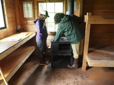 Two people lighting the stove in Gut Island Cabin 2 with wooden bunkbeds and cabinets