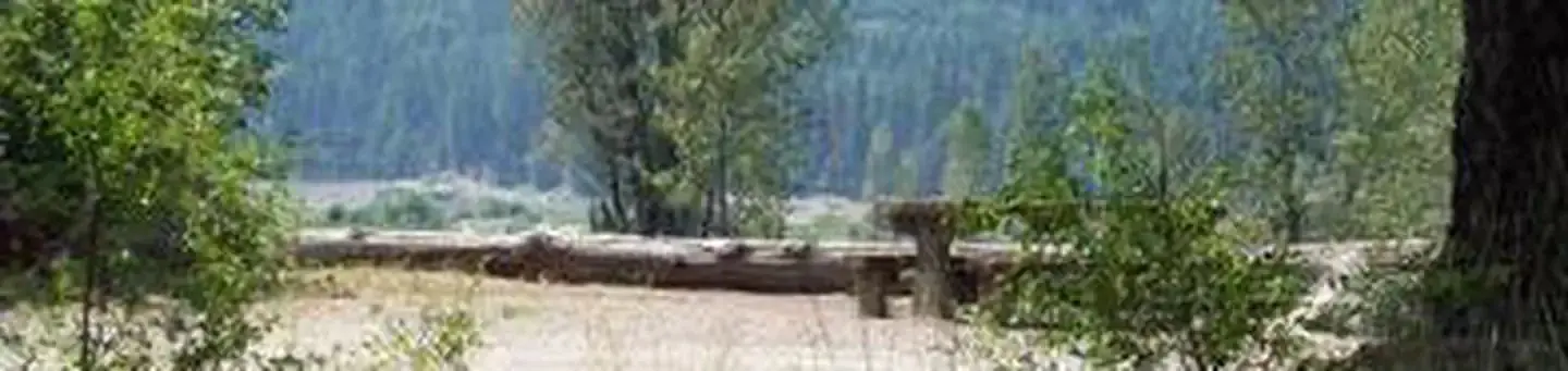 Picnic table in a flat gravel area with bushes and log parking barrier, river valley and conifer covered hills in background.