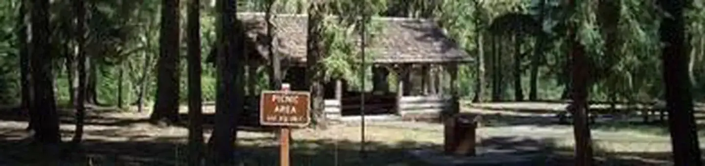 Picnic area sign in foreground,  sunlit group picnic shelter set in conifer forest background.