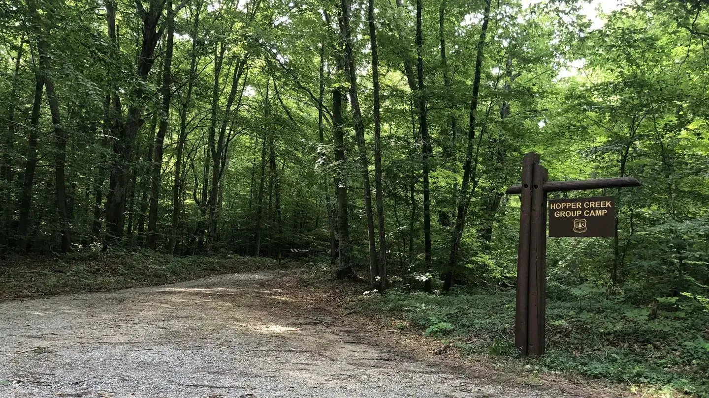 Entrance drive to Hopper Creek Group Camp, a gray gravel driveway is on the left and a brown sign indicating Hopper Creek Group Camp is on the right.
