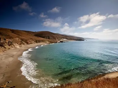 Sandy beach with ocean and small waves and steep coastal bluffs covered in dry grass.