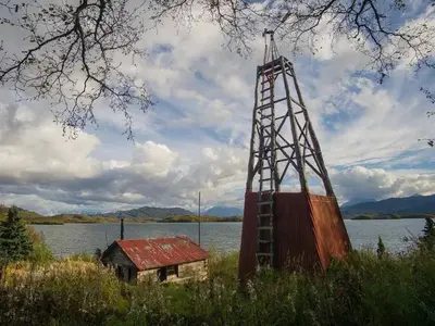 View of a wood cabin and windmill in a grassy field with a lake and mountains in the background.