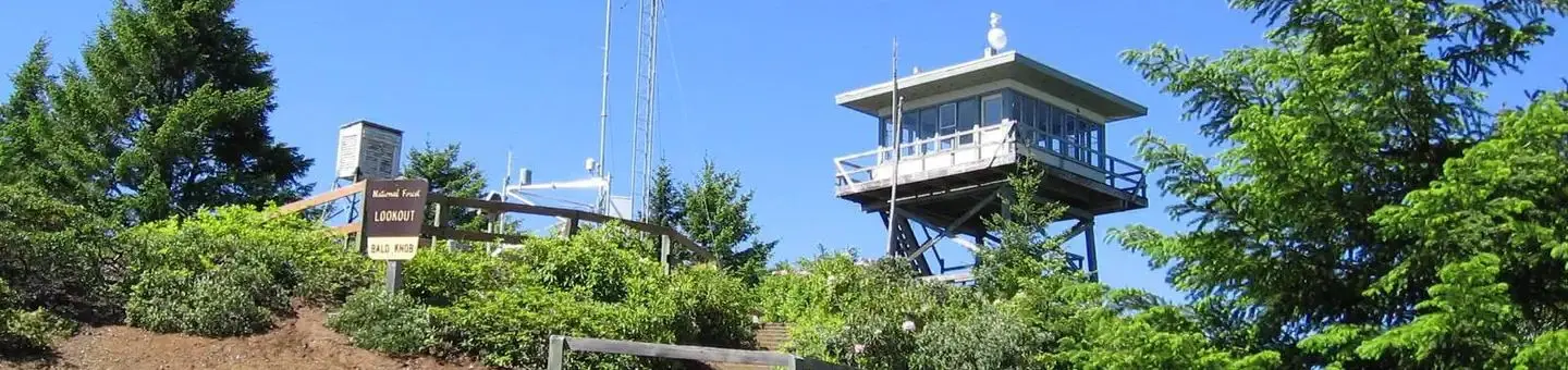 Lookout building on ridge with Forest Service i.d. sign, communications tower, green shrubs and conifer trees backed by blue sky.