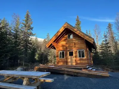 Two-story log cabin with large front porch and ramp. A picnic table is in the foreground.