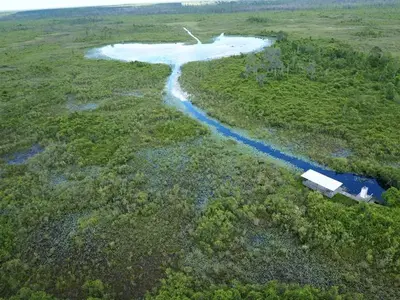 Aerial view of a round open lake in the background with a narrow trail into the foreground leading to a shelter