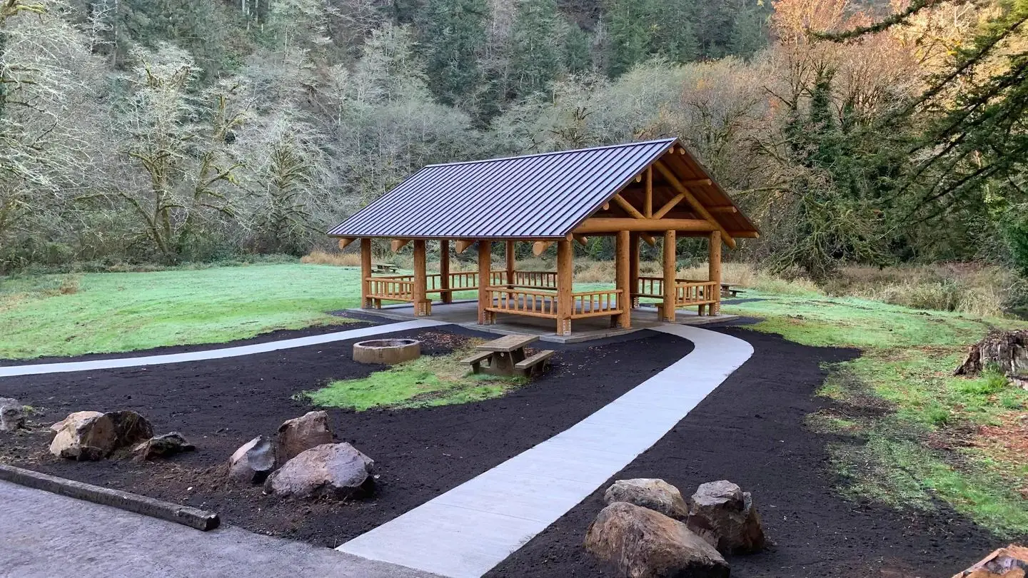 Picnic shelter.  Large flat field.  Tents and picnic tables in front of a bridge, a large conifer tree, and mixed conifer forest in the back, fire ring and picnic table behind single pole fence in foreground.