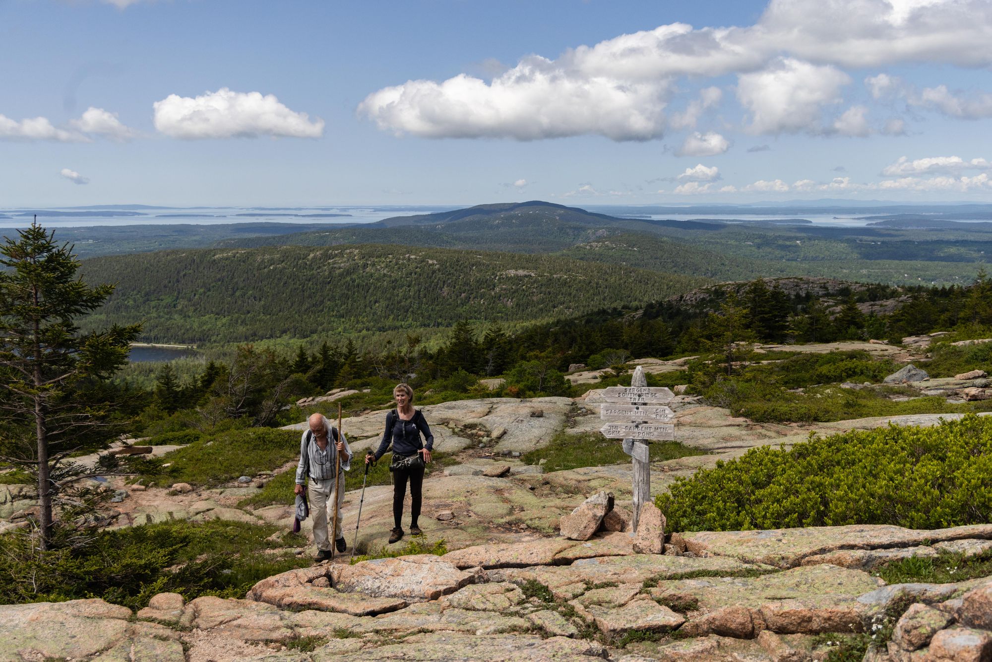 A man and woman holding walking sticks hike up a mountain summit near a wooden trail sign.