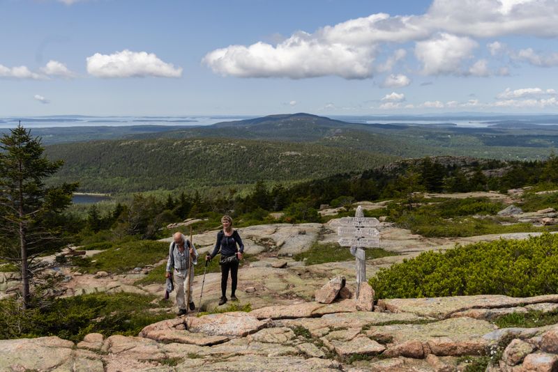 A man and woman holding walking sticks hike up a mountain summit near a wooden trail sign.