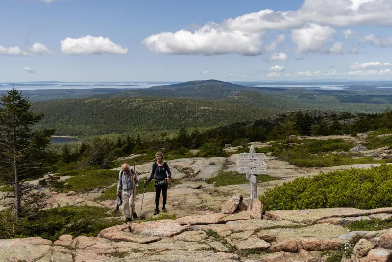 A man and woman holding walking sticks hike up a mountain summit near a wooden trail sign.