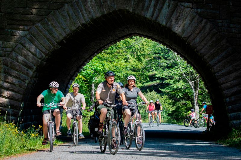 A group of people riding bicycles travels under the arch of a large stone bridge