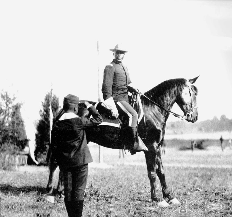 An African American man in a military uniform sitting high on a horse being saluted by a boy