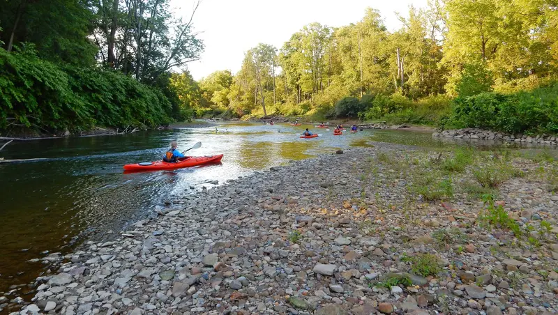 People in bright orange kayaks paddle around a bend in a river, past green trees and a rocky shore.