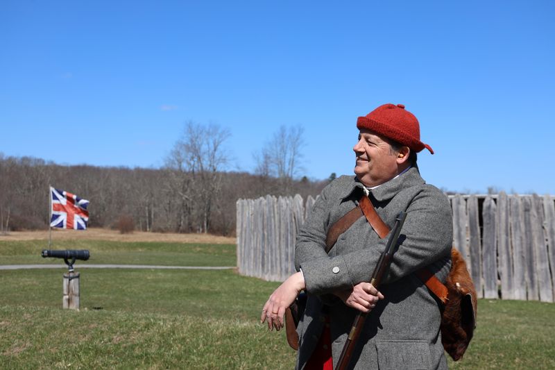Man in 18th century flintlock musket stands in front of a round stockade fort with a British flag