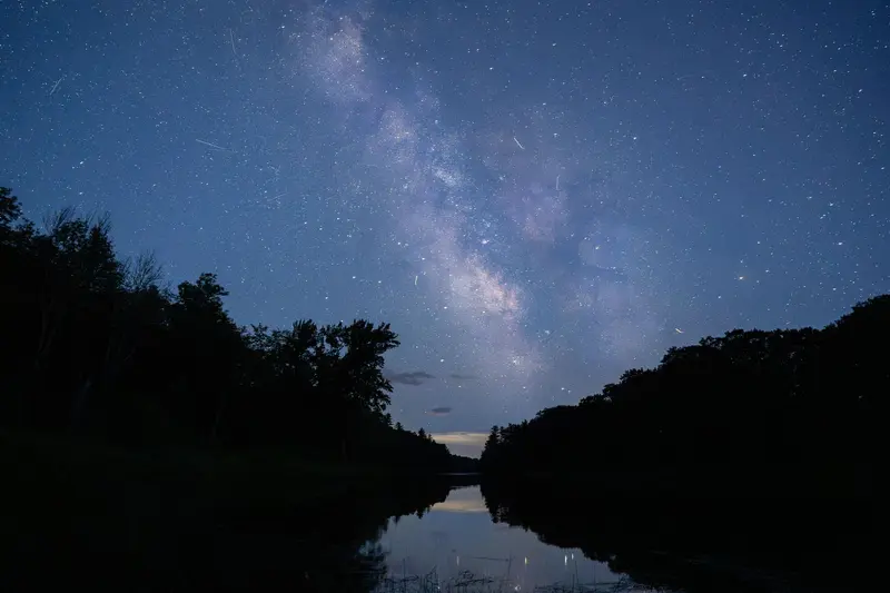 A blue night sky filled with stars sits over reflective water. Dark trees line the image.