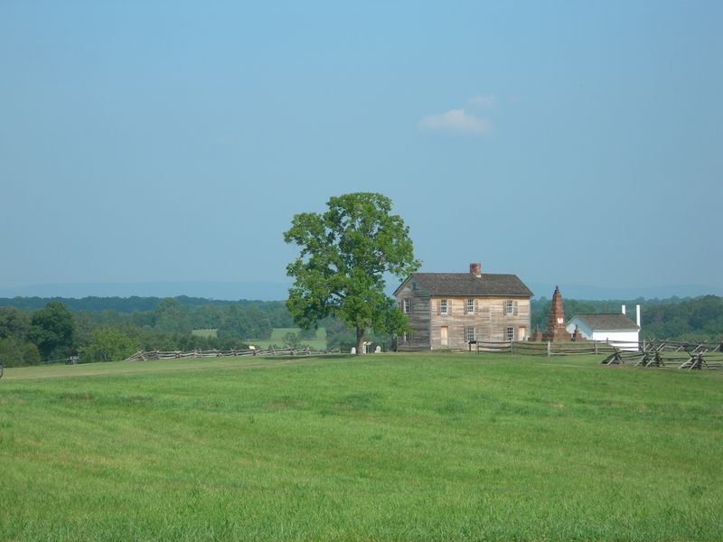 Rustic farmhouse, flanked by shade tree, stone monument, and white outbuilding, on top of a hill.