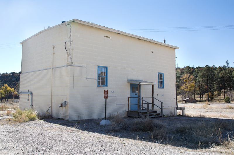 Two-story white house with two small windows, a single door, and flat roof stands in a gravel lot.
