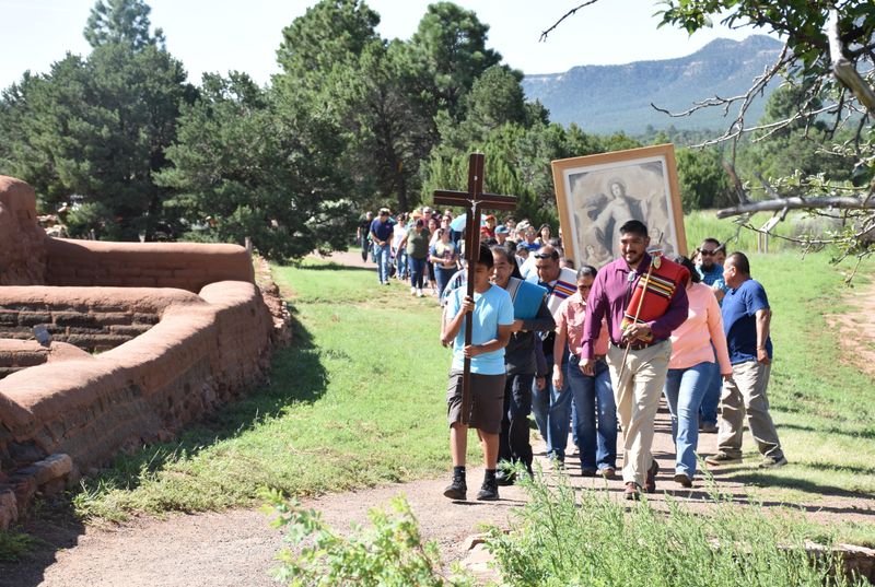 A line of people walk past adobe bricks and trees on a path