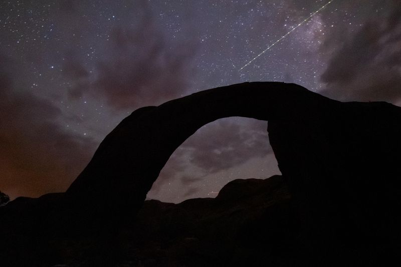 A silhouetted rock arch under a stary sky with a satellite flying by.