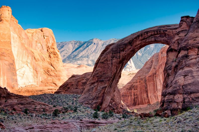 A natural stone arch sits between red rock canyon walls.  A mountain is seen through the arch.
