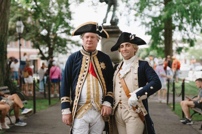 Two reenactors in Revolutionary War-era military uniforms stand together in a park.